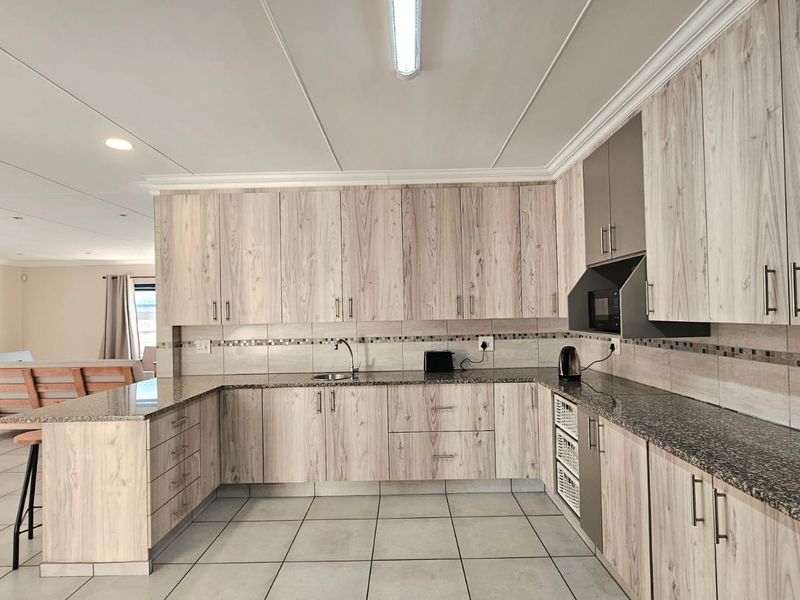 Spacious open-plan kitchen featuring light wood-grain cabinetry, granite countertops, and tiled flooring, seamlessly flowing into the adjacent living area.