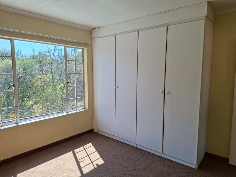 A bright, empty bedroom featuring built-in cupboards, carpeted flooring, and a large window with a view of the surrounding trees.
