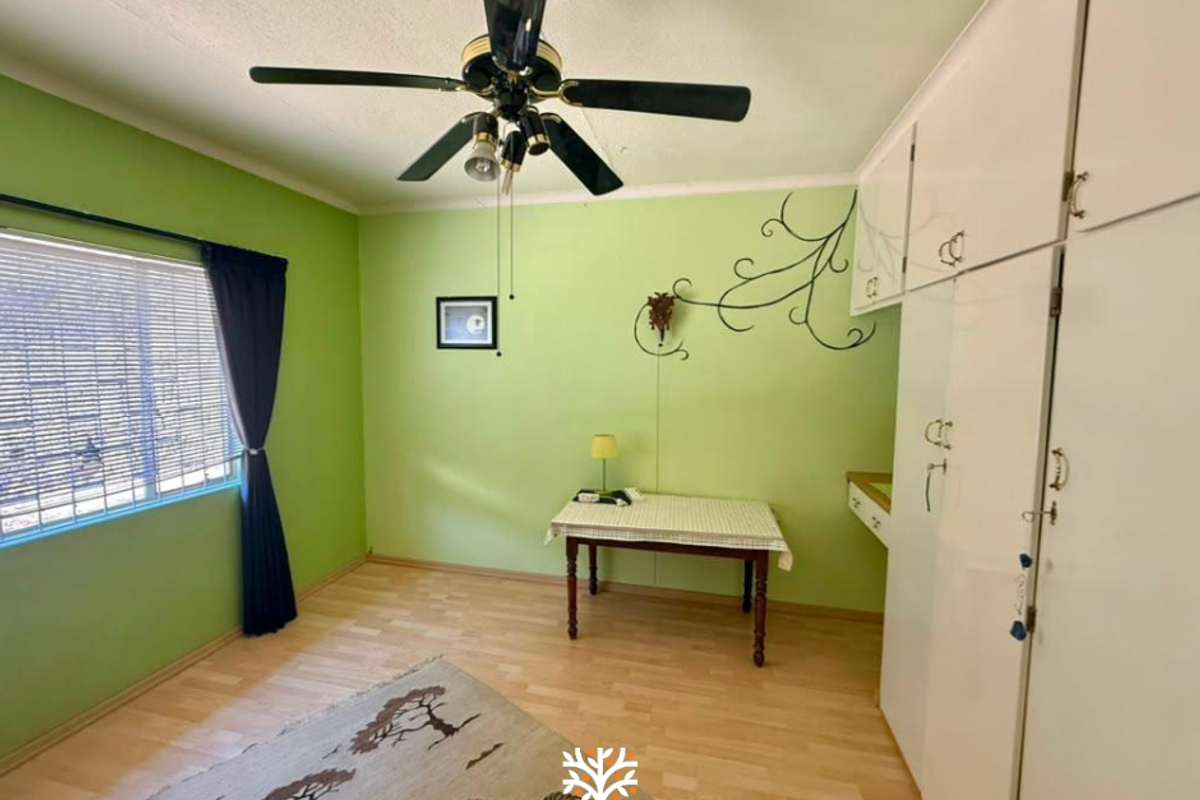 A bedroom featuring laminate flooring, built-in cupboards, and a ceiling fan, with large windows fitted with venetian blinds.