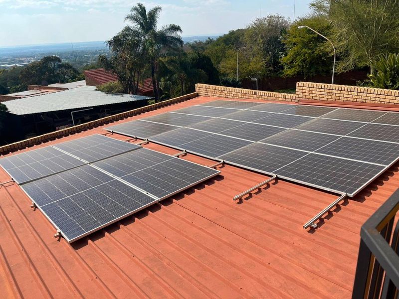 A view of a metal roof featuring a partially installed solar panel array with a brick parapet wall and surrounding landscape.