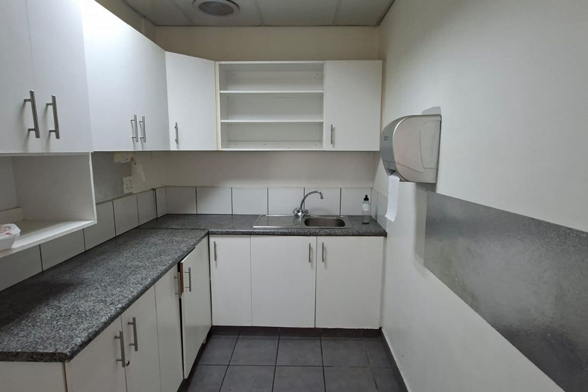 A functional kitchenette area featuring white cabinetry, granite-look countertops, a stainless steel sink, and tiled flooring, suitable for office or industrial utility use.