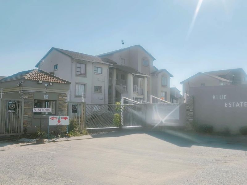 Front entrance view of the apartment complex featuring a security gate, guard house, and paved driveway access.