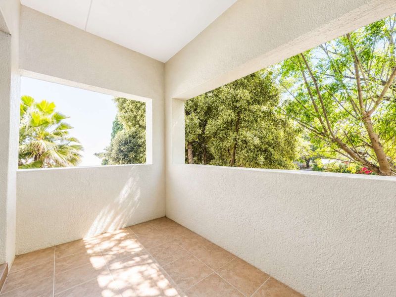 A clean, covered balcony featuring neutral tiled flooring and plastered walls, offering an open view of the surrounding greenery.
