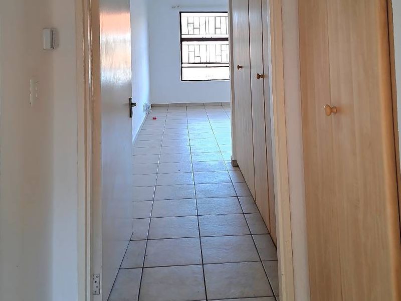 A functional hallway featuring tiled flooring and built-in storage cupboards, leading towards a bright bedroom area with security bars on the window.
