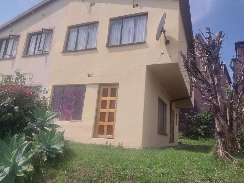 Rear view of a two-story residential property featuring a plastered facade, timber entrance door, and multiple casement windows set within a garden area.