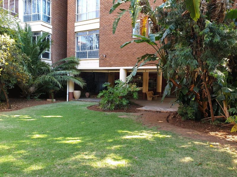 A view of the apartment building's rear facade featuring a covered patio area overlooking a well-maintained garden with mature landscaping.