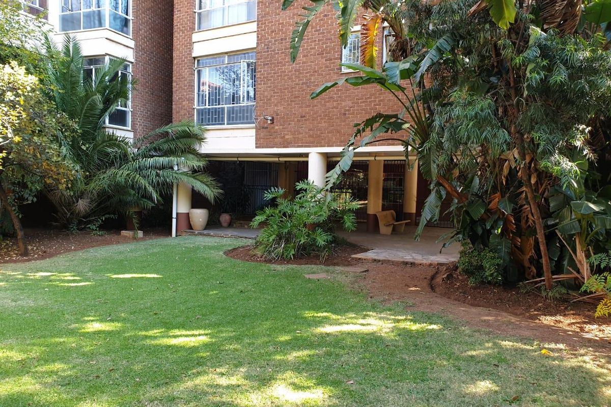 A view of the apartment building's rear facade featuring a covered patio area overlooking a well-maintained garden with mature landscaping.