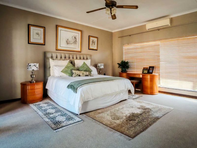 Spacious bedroom featuring neutral wall tones, carpeted flooring, a ceiling fan, and a wall-mounted air conditioning unit with large windows and venetian blinds.