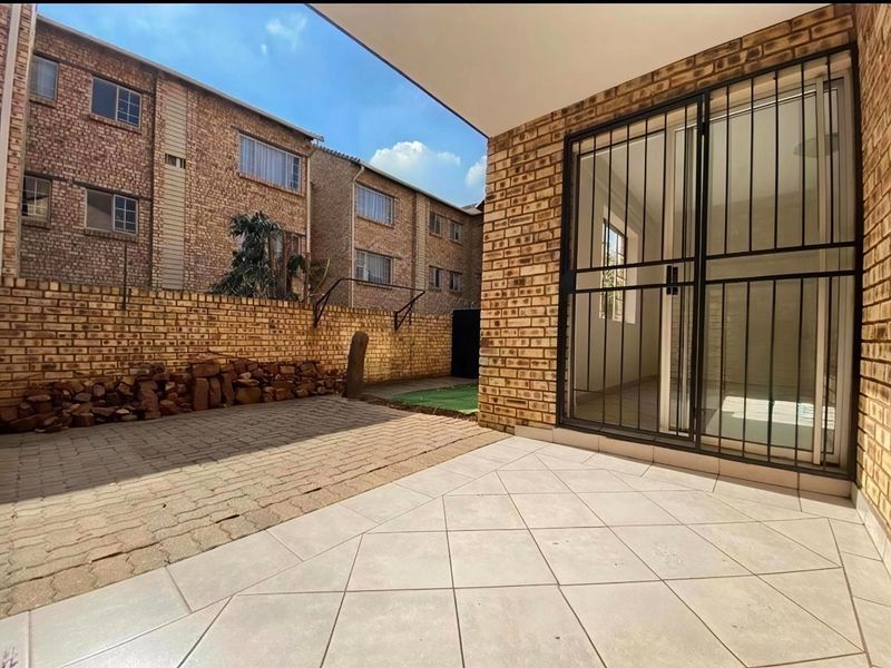 A private paved patio area featuring a security gate, sliding glass door access, and a brick boundary wall in a residential apartment complex.