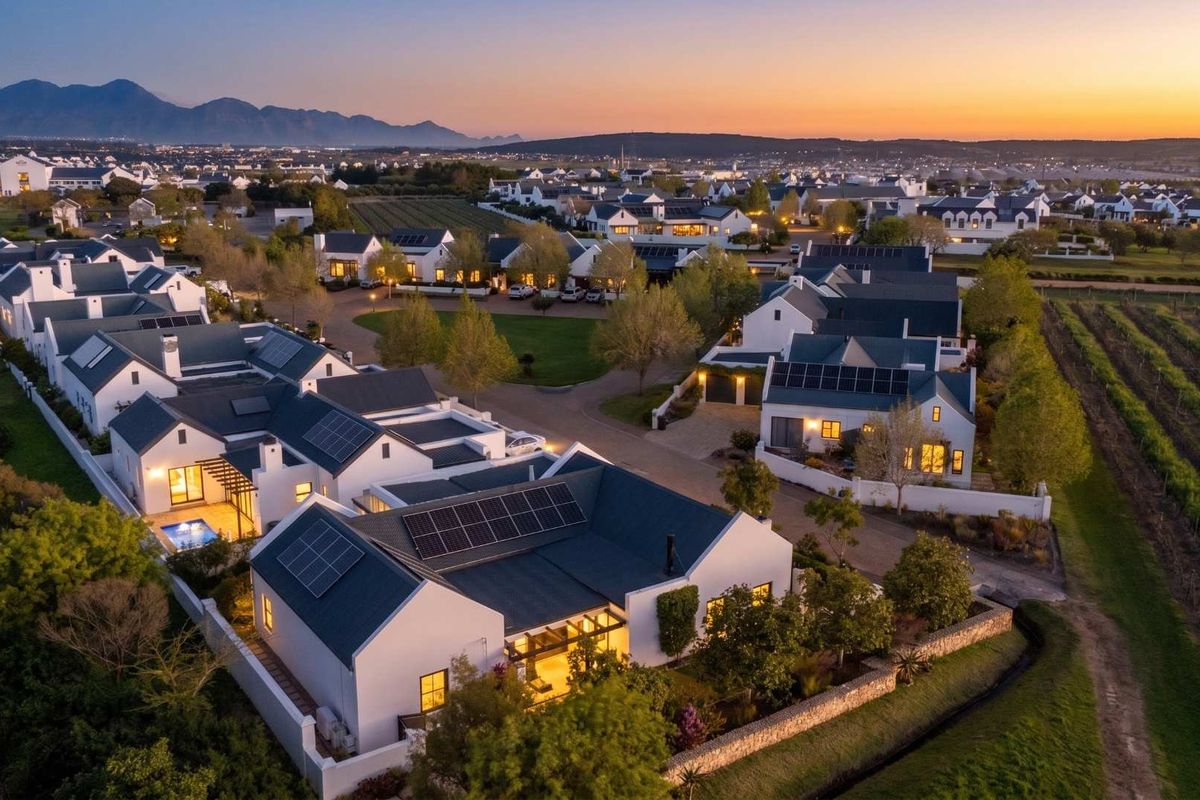 An elevated aerial view of a modern residential estate at sunset, showcasing multiple homes with solar panels, manicured gardens, and a swimming pool.
