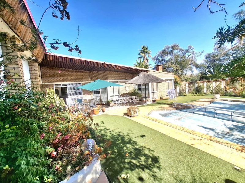 Rear exterior view featuring a swimming pool, covered patio area, and established garden with brick facade and security gates.