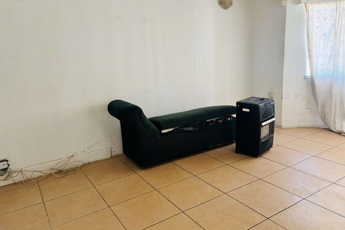 A sparsely furnished living area featuring ceramic tiled flooring and wall-mounted lighting, showing signs of wear on the walls and skirting boards.