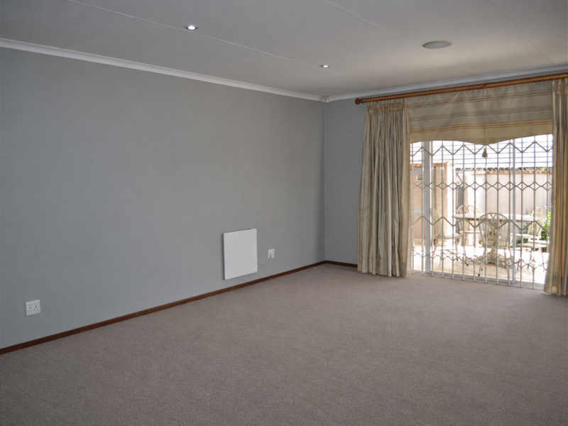 A spacious, neutral-toned living room featuring carpet flooring, recessed lighting, and a sliding glass door with security gate leading to an outdoor patio area.