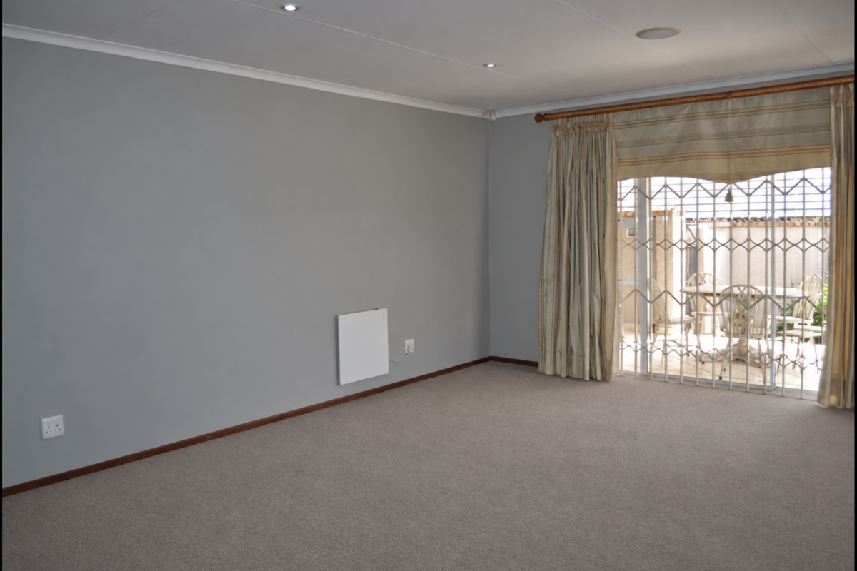 A spacious, neutral-toned living room featuring carpet flooring, recessed lighting, and a sliding glass door with security gate leading to an outdoor patio area.