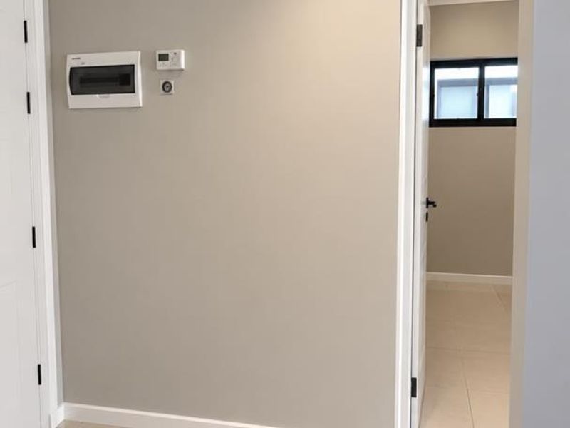 A clean, modern hallway featuring neutral wall tones, large format floor tiles, and integrated electrical fixtures, leading into an adjacent room.