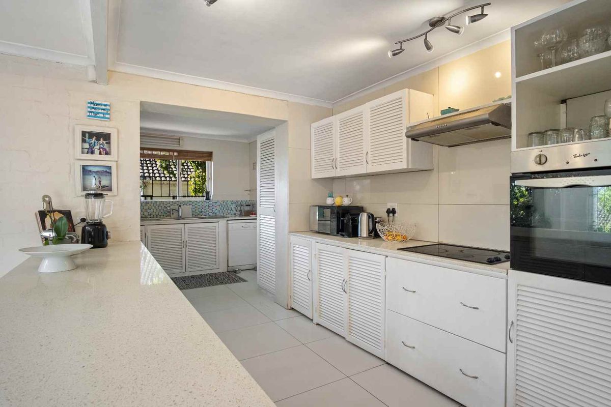 A functional kitchen featuring white louvered cabinetry, stone countertops, integrated oven and hob, and a separate scullery area with tiled flooring.