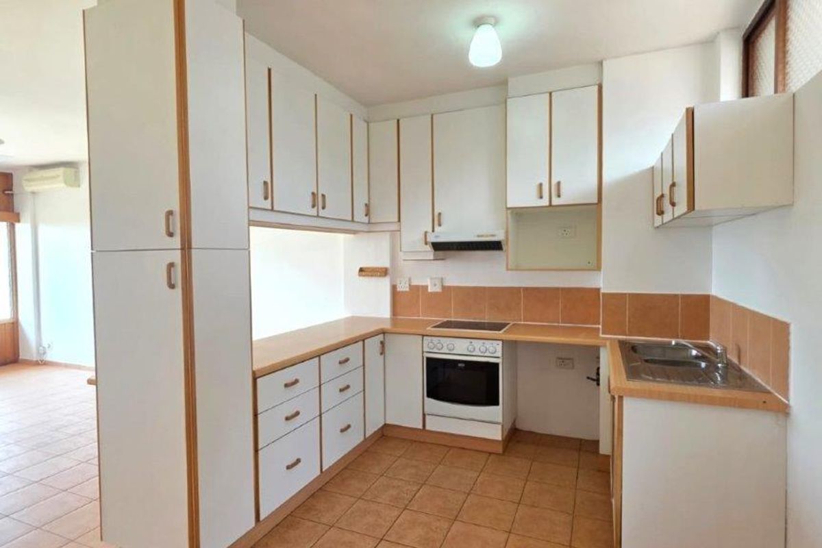 A functional kitchen featuring white cabinetry, laminate countertops, integrated oven and hob, and tiled backsplash, opening into an adjacent living area.