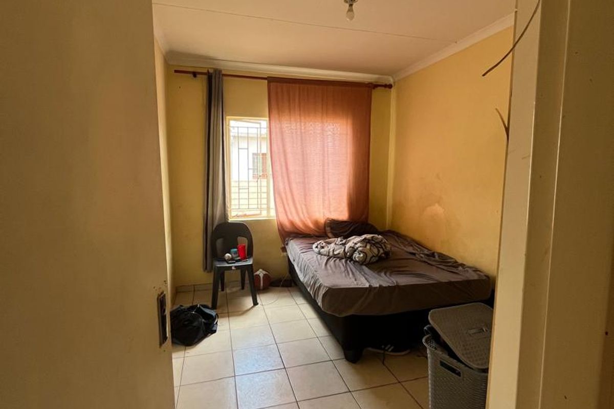 A compact bedroom featuring tiled flooring, a window with security bars, and basic lighting fixtures, requiring cosmetic updates to improve its overall condition.
