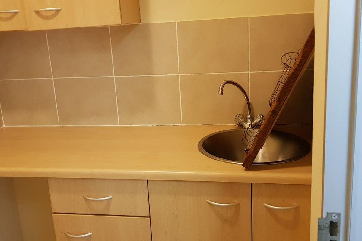 A compact kitchenette or utility area featuring light wood-finish cabinetry, a stainless steel sink, and neutral tiled splashback.