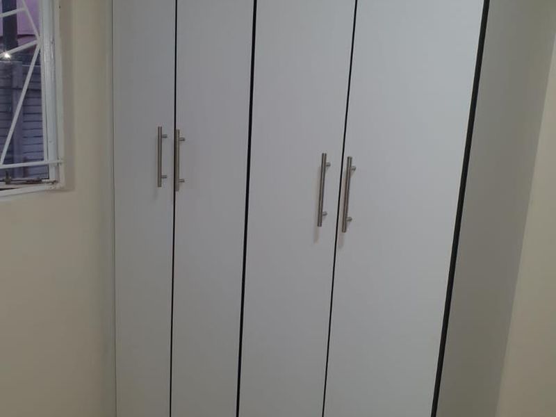 A clean bedroom interior featuring modern white built-in cupboards, tiled flooring, and a window fitted with security bars.