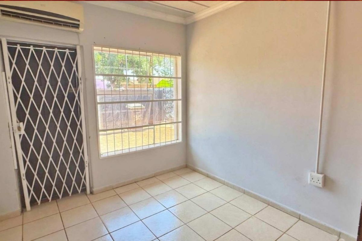 A functional, empty room featuring tiled flooring, a security gate, a window with security bars, and a wall-mounted air conditioning unit.
