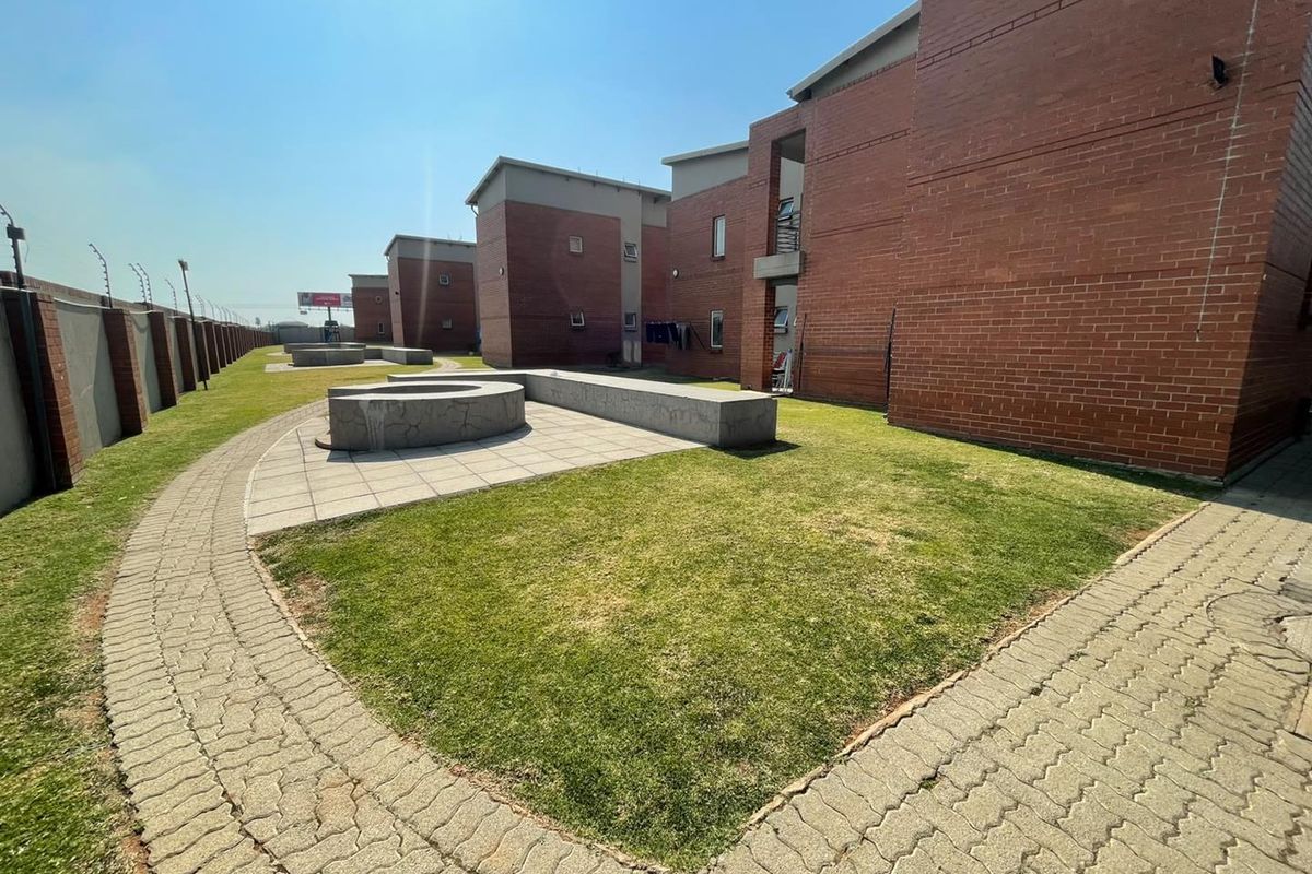 A view of the rear exterior of the apartment complex, featuring brick facades, paved walkways, and a communal lawn area with concrete seating.
