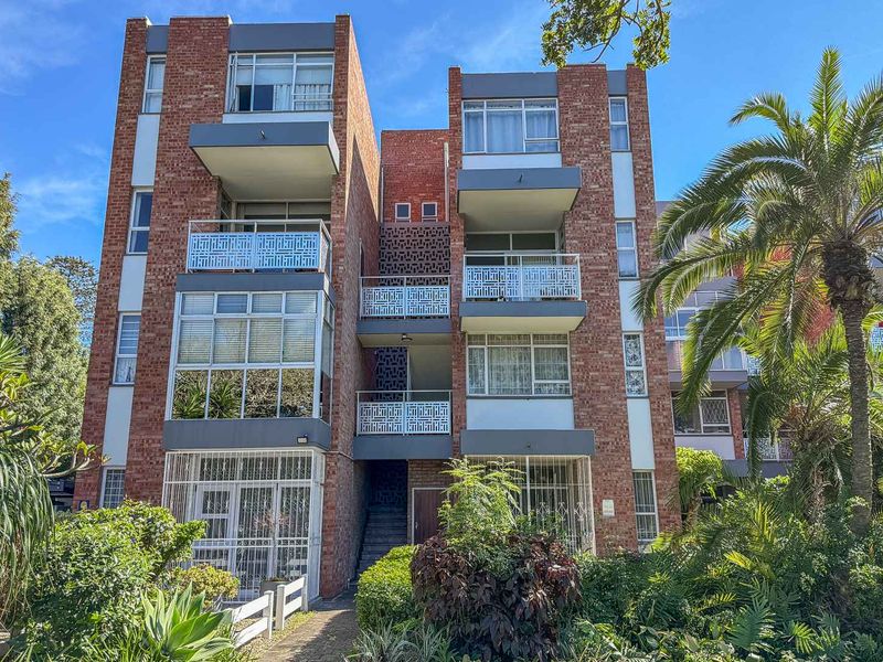 Exterior view of a multi-story brick apartment building featuring private balconies with metal railings and established garden surroundings.