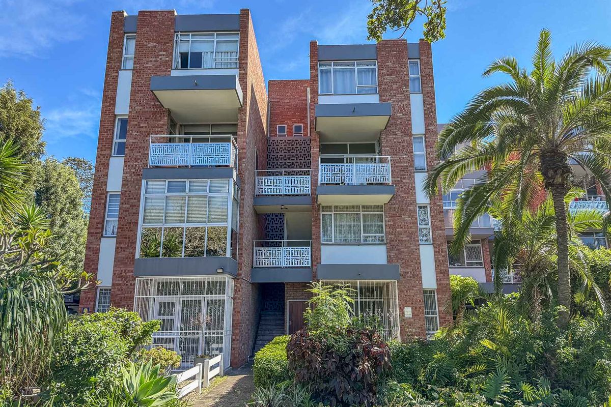 Exterior view of a multi-story brick apartment building featuring private balconies with metal railings and established garden surroundings.