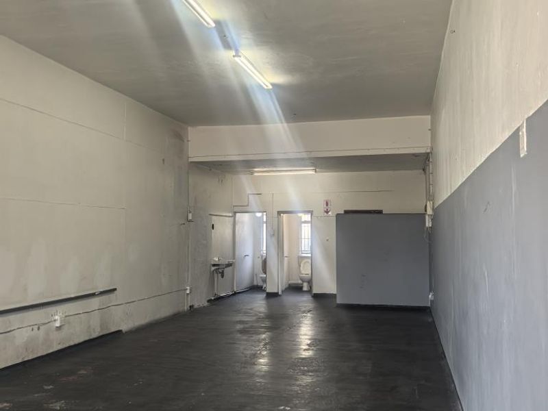 An empty, utilitarian commercial space featuring concrete flooring, fluorescent lighting, and a small rear area with a basin and toilet.