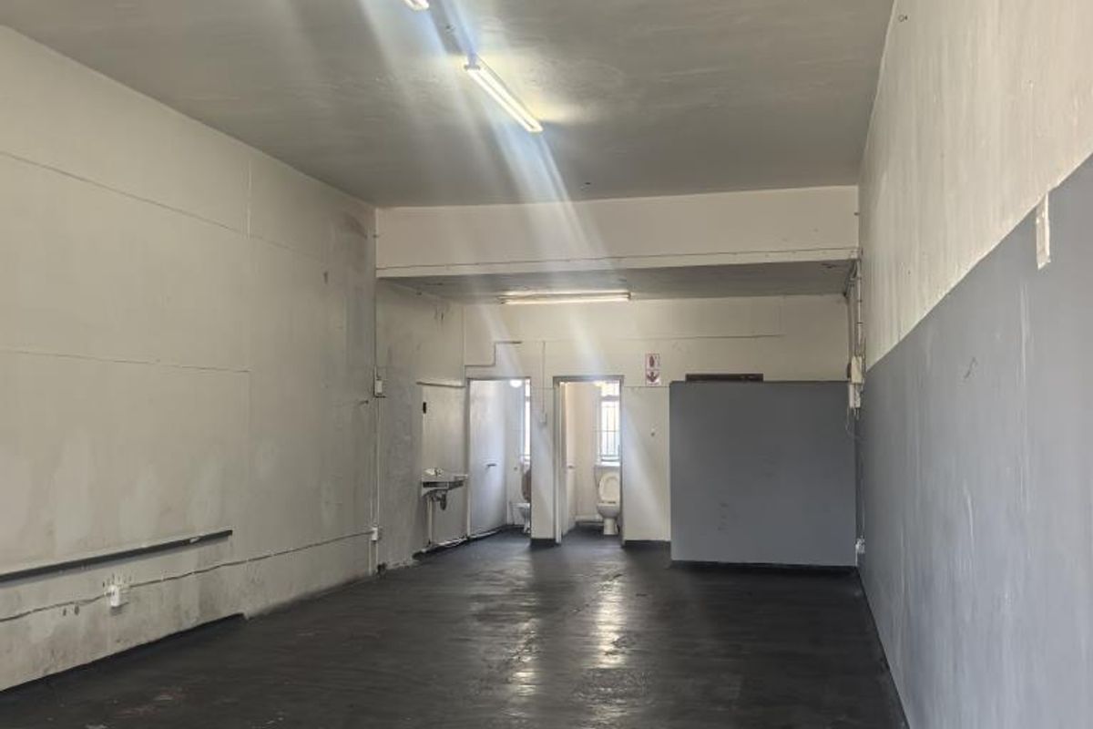 An empty, utilitarian commercial space featuring concrete flooring, fluorescent lighting, and a small rear area with a basin and toilet.