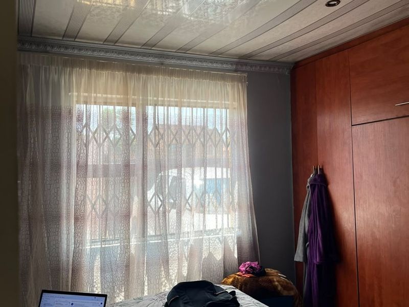 A bedroom featuring built-in wooden cupboards, decorative ceiling panels with recessed lighting, and a window fitted with a security gate.