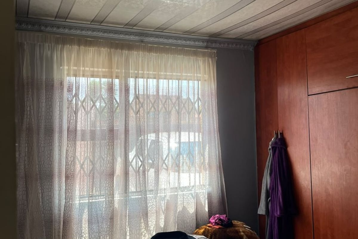 A bedroom featuring built-in wooden cupboards, decorative ceiling panels with recessed lighting, and a window fitted with a security gate.