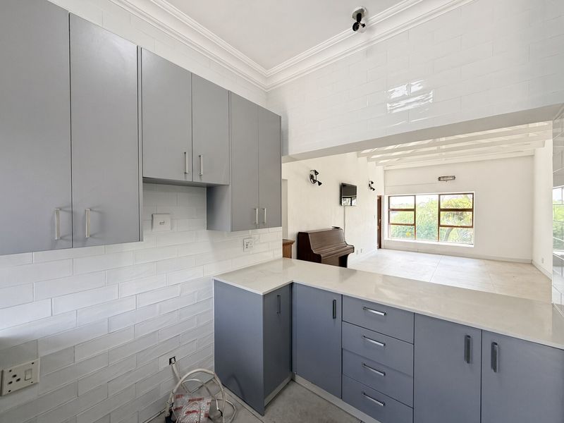 Modern open-plan kitchen featuring sleek grey cabinetry, stone countertops, and white subway tile backsplash, flowing into a bright, spacious living area with large windows.