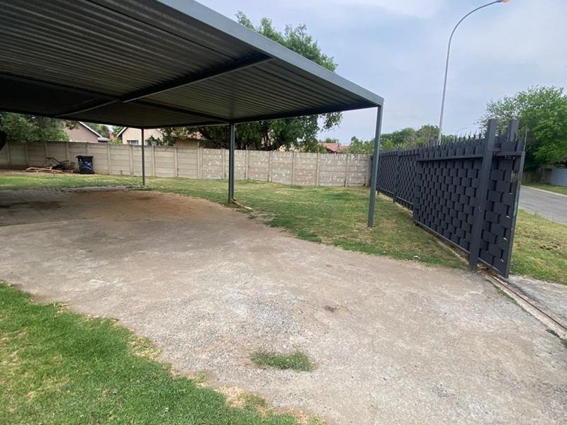 Outdoor carport area featuring a metal roof structure, concrete driveway, and secure perimeter fencing with a metal gate.
