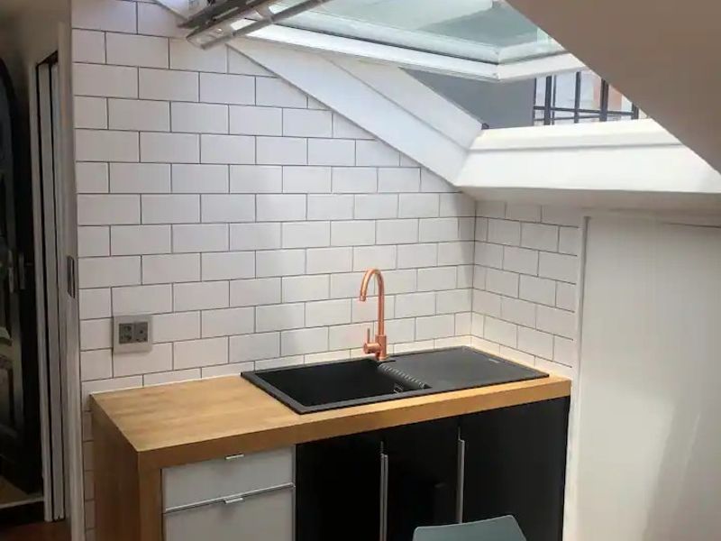 A compact kitchen area featuring a modern black sink, copper faucet, wooden countertop, and subway tile backsplash, illuminated by a large skylight.