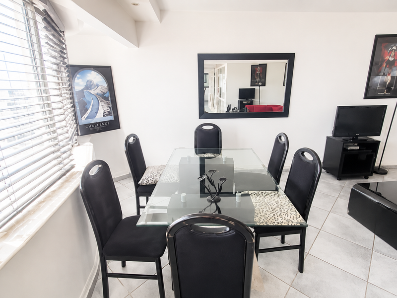 A bright living and dining area featuring neutral tiled flooring, large windows with venetian blinds, and recessed ceiling lighting.