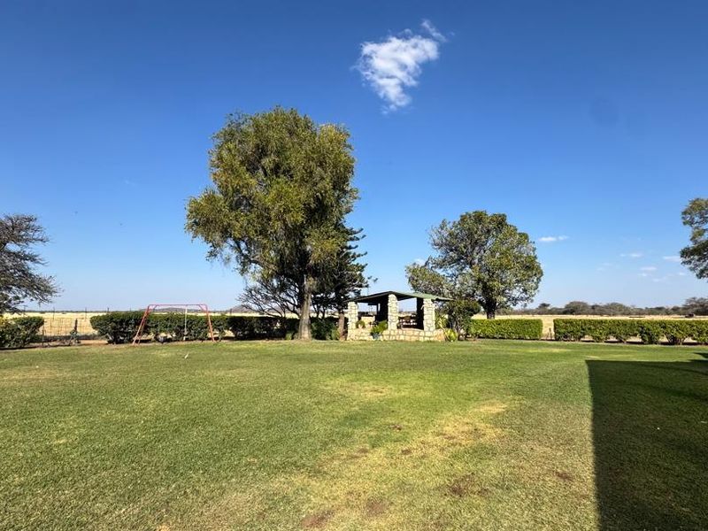 A spacious, well-maintained farm garden featuring a stone gazebo, mature trees, and neatly trimmed hedging under a clear blue sky.