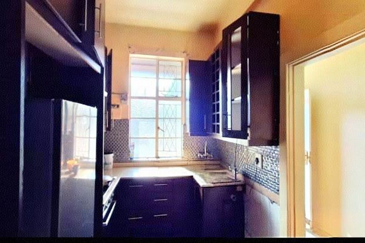 A compact kitchen featuring dark wood cabinetry, mosaic tile backsplash, and a window with security bars, requiring minor finishing work to the lower wall area.