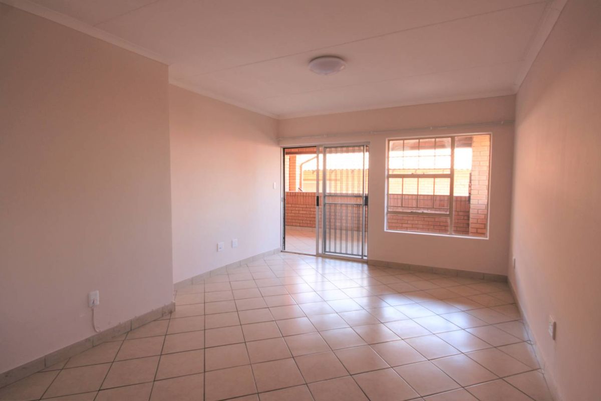 A bright, empty living room featuring neutral tiled flooring, a sliding glass door leading to a balcony, and large windows for natural light.