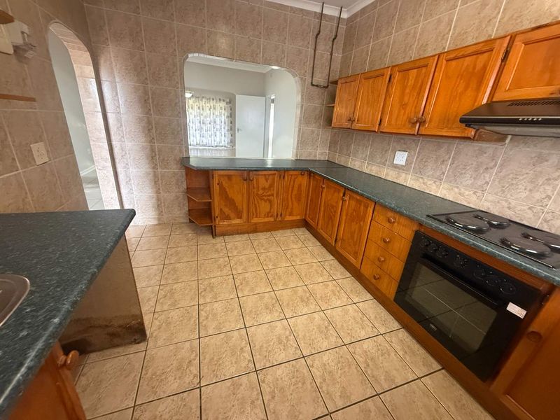 A dated kitchen featuring wooden cabinetry, laminate countertops, and ceramic tiling, offering a functional layout with an open-plan archway to the adjacent living area.