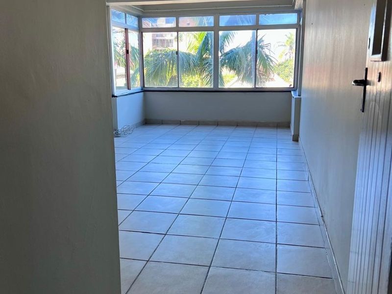 An empty, sunlit living area featuring neutral tiled flooring, a ceiling fan, and large windows providing natural light and a pleasant outlook.