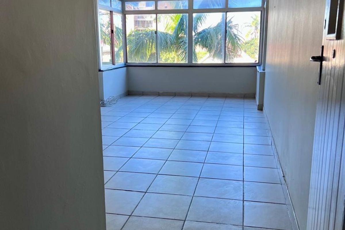 An empty, sunlit living area featuring neutral tiled flooring, a ceiling fan, and large windows providing natural light and a pleasant outlook.