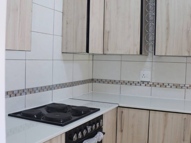 A clean, modern kitchen featuring light wood-finish cabinetry, a built-in electric oven and hob, and neutral tiled backsplash and flooring.