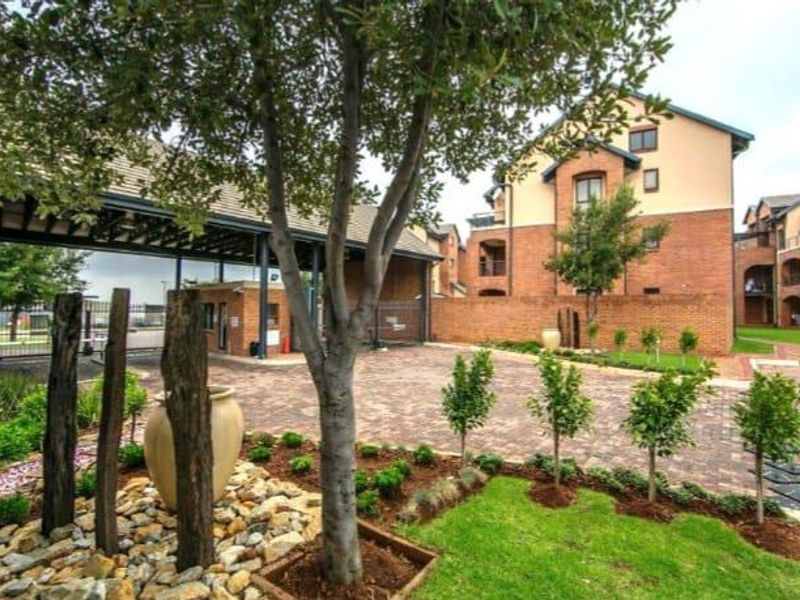 A well-maintained apartment complex entrance featuring brick paving, landscaped garden beds, and a secure gated access point.