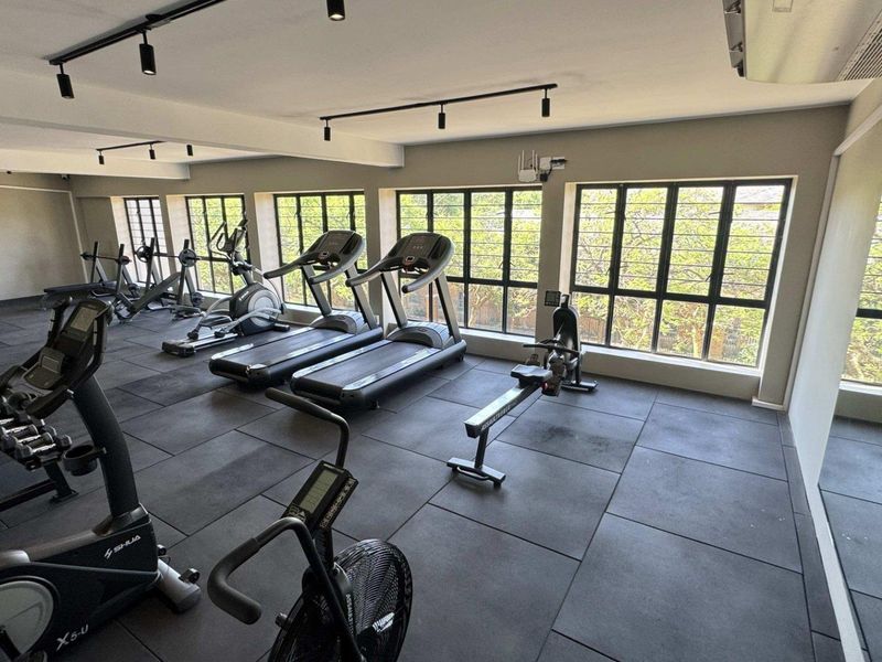 A well-lit, spacious gym area featuring durable rubber flooring, track lighting, and large windows providing natural light.