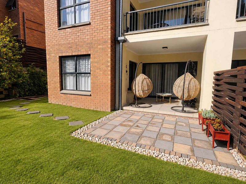 A well-maintained rear exterior featuring a paved patio area, a small lawn, and a timber privacy screen, adjacent to the apartment building.