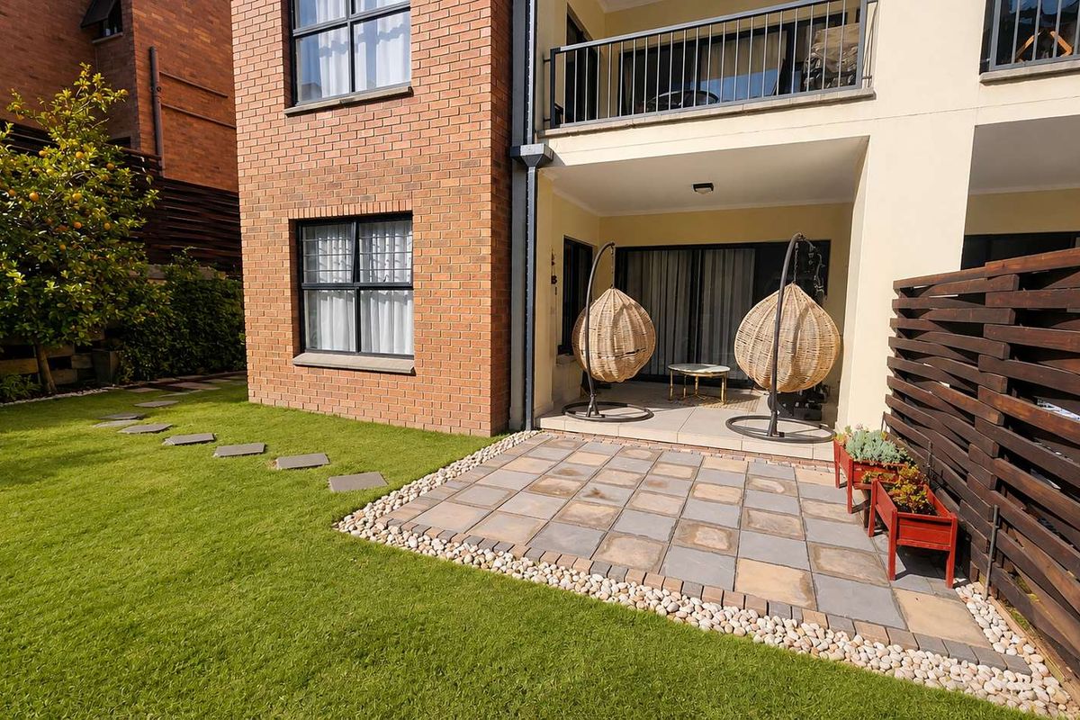A well-maintained rear exterior featuring a paved patio area, a small lawn, and a timber privacy screen, adjacent to the apartment building.