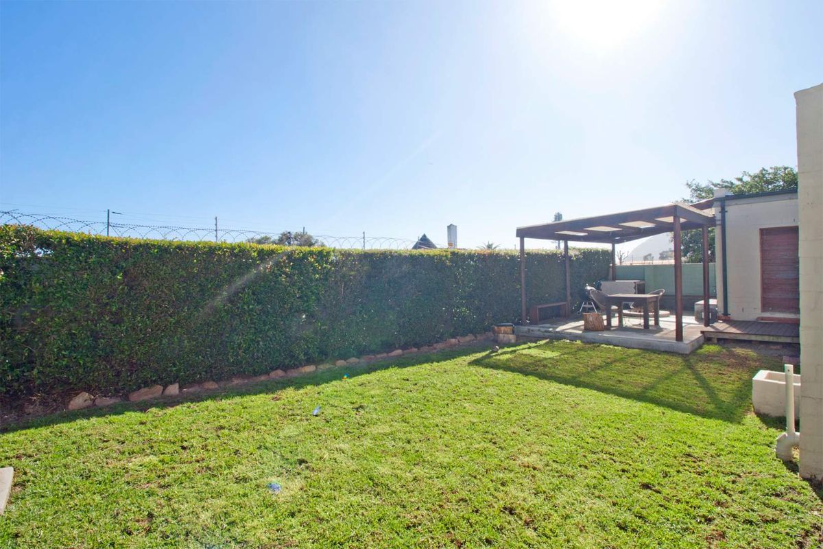 A private rear garden featuring a well-maintained lawn, a timber pergola, and a raised timber deck area enclosed by a tall hedge.
