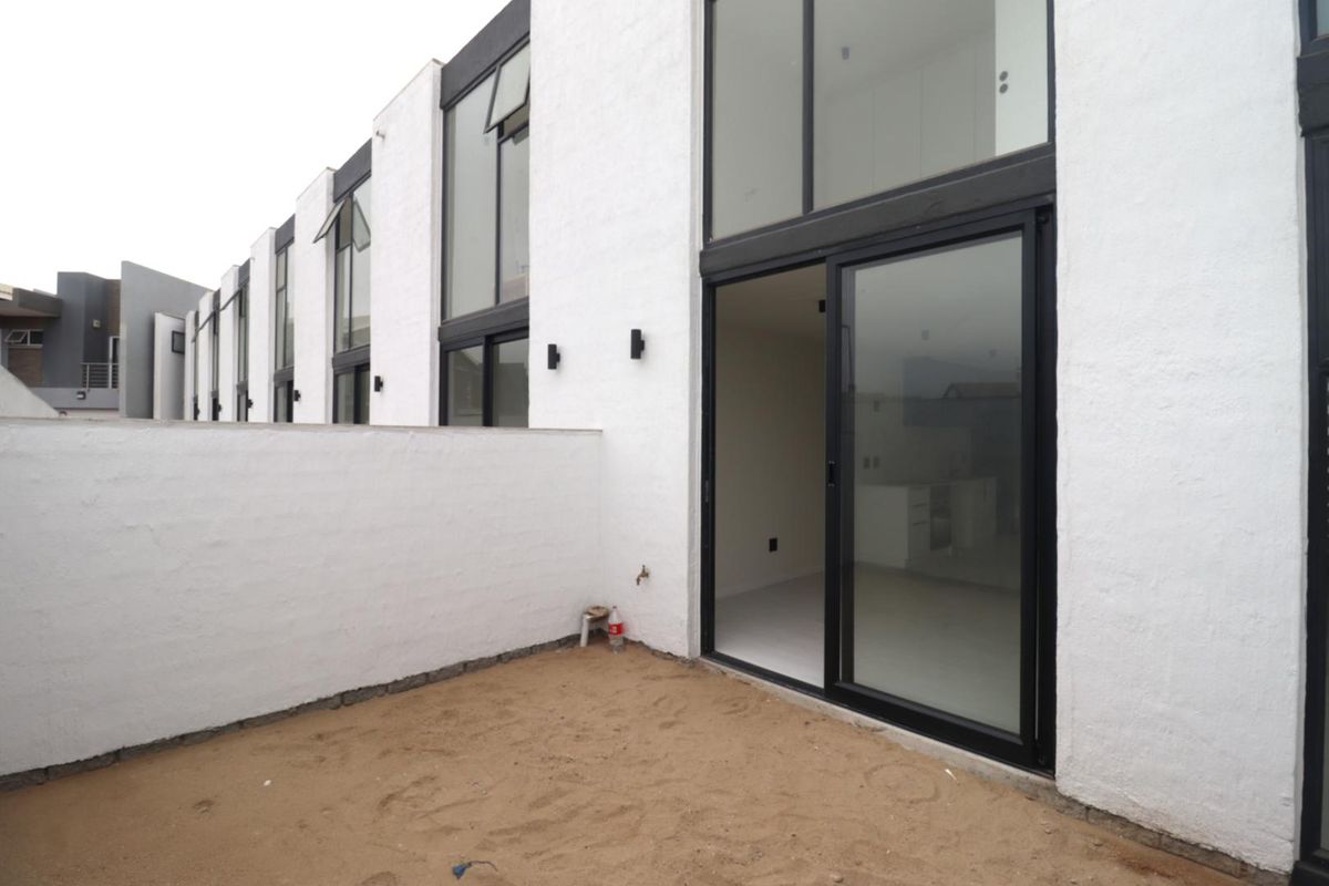 Rear view of a modern apartment complex featuring white textured facades, large glass sliding doors, and an unfinished ground area.