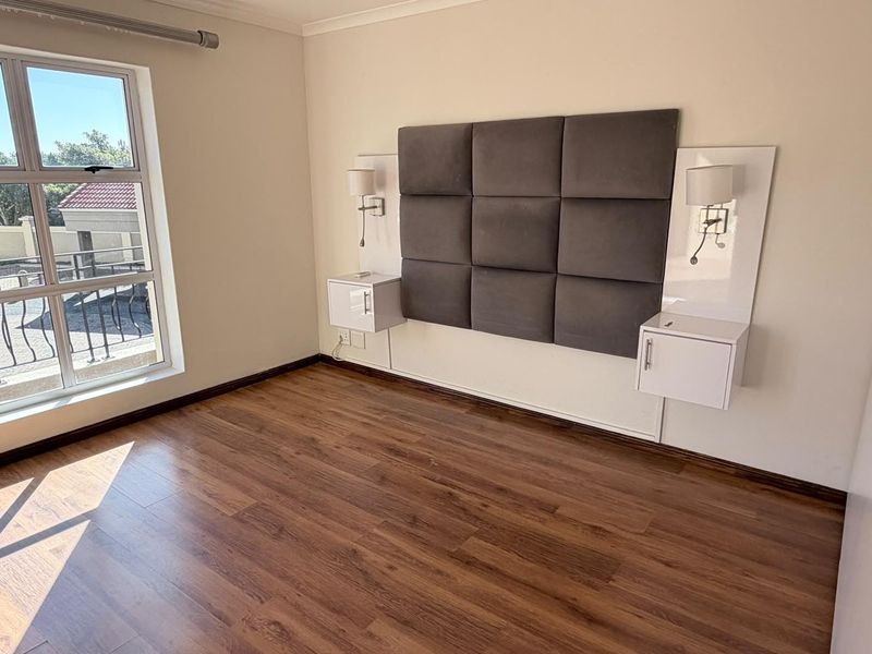 A bright, empty bedroom featuring laminate flooring, a modern upholstered headboard, and integrated wall-mounted bedside cabinetry with built-in lighting.